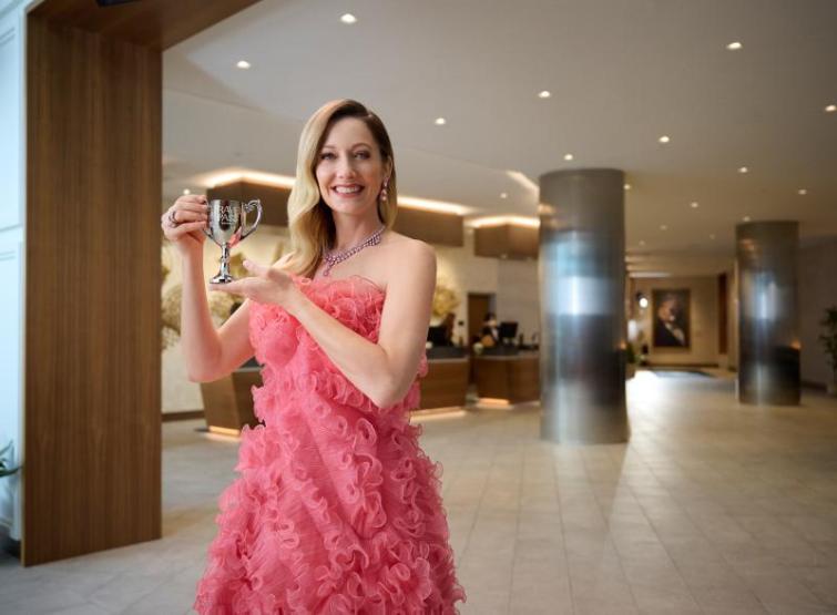 Judy Green in stylish Pink dress holding up Travel Pass Rewards Trophy in beautiful hotel lobby